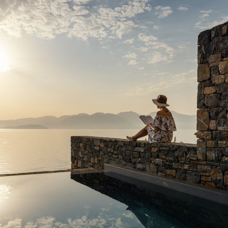 Frau sitzt nebem dem Pool auf einer Mauer und liest ein Buch. Sie blickt auf das weite Meer
