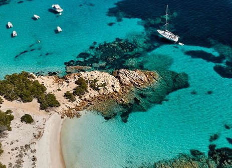 Strand in Sardinien mit Booten im Wasser und einer felsigen Küste im Hintergrund.