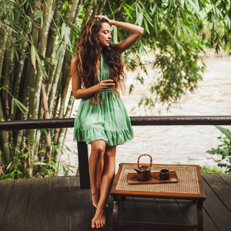 Frau in grünem Rock und langen, braunen Haaren steht mit einer Tasse Tee in der Hand auf einer Terrasse. Im Hintergrund fliesst ein Fluss