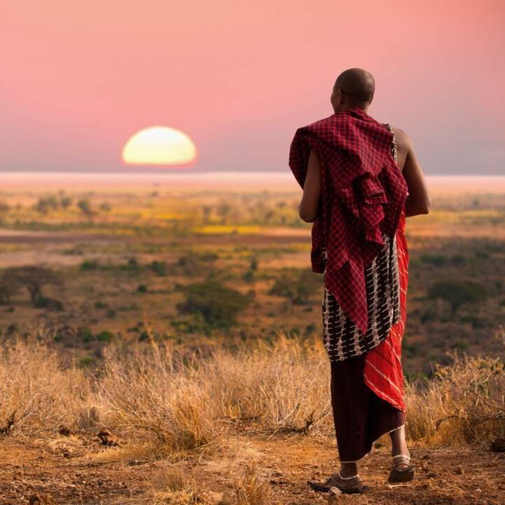 Ein Mann in einem roten Gewand steht bei Sonnenuntergang auf einer Ebene in Tansania.