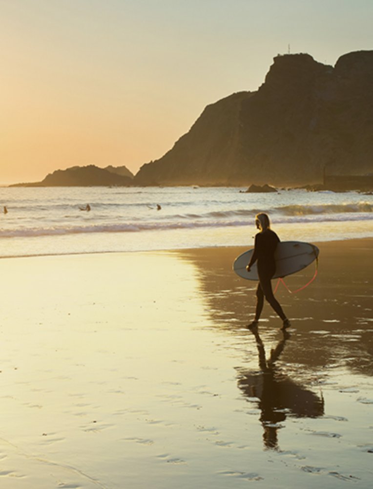 Une personne marche sur la plage de l'Algarve avec une planche de surf sous le bras.