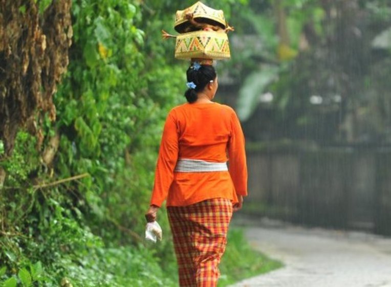 Balinese woman wearing an orange sarong while carrying a fruit basket on her head down a road