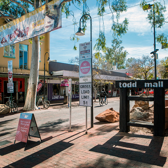 Zone piétonne de la Todd Mall à Alice Springs avec panneau en bois, arbres offrant de l’ombre, boutiques, vélos et passants le long de l’allée pavée.