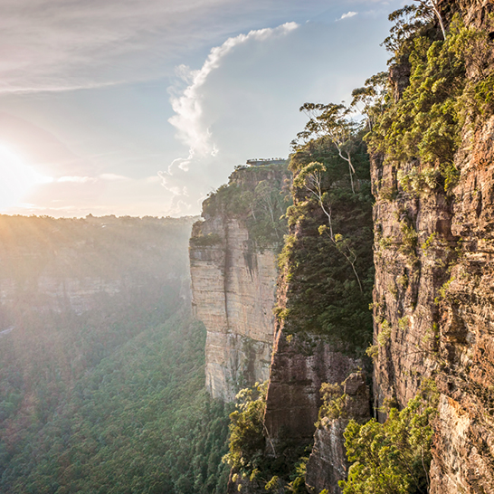 Falaises abruptes des Blue Mountains avec une végétation verdoyante, une vallée boisée en contrebas et une lumière chaude filtrant à travers un ciel partiellement nuageux.