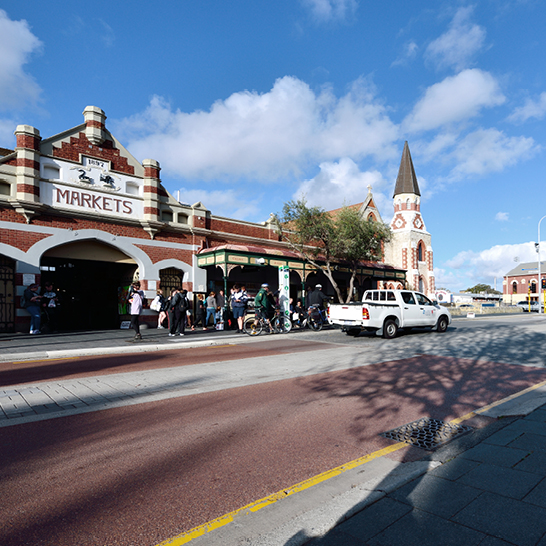Scène de rue devant le bâtiment historique en briques des Fremantle Markets, avec entrée voûtée, passants, vélos et un pick-up garé le long de l’allée pavée.