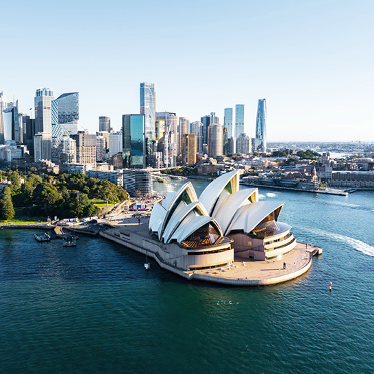 Vue sur l’Opéra de Sydney avec ses toits blancs en forme de voiles, situé au bord du port, entouré d’eaux bleues et de la skyline de Sydney en arrière‑plan.