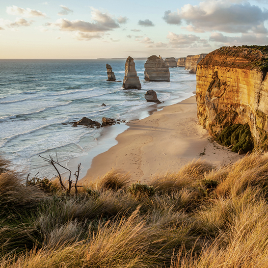 Paysage côtier avec les hautes formations calcaires des Twelve Apostles, des falaises abruptes, une plage de sable, des vagues douces et un ciel partiellement nuageux baigné d’une lumière chaude.