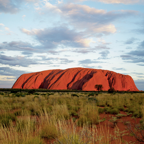 Vue aérienne d’Uluru, le vaste monolithe de grès rouge‑orangé au cœur du désert australien, entouré d’un paysage plat à végétation clairsemée sous un ciel nuageux.