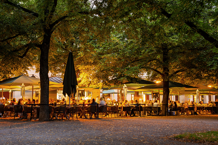 Beleuchteter Biergarten in Deutschland am Abend, mit vielen Gästen unter grossen Bäumen und grünen Sonnenschirmen.
