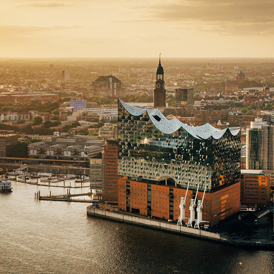 Elbphilharmonie im Hamburger Hafen bei Sonnenuntergang, mit leuchtender Glasfassade und Blick auf die umliegende Stadt.