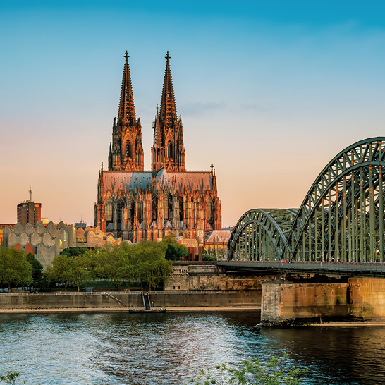 Kölner Dom und Hohenzollernbrücke am Rhein im warmen Abendlicht, mit Spiegelungen auf dem Wasser.