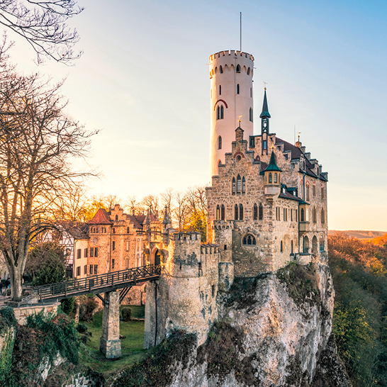Schloss Neuschwanstein auf einem Felsen in Baden‑Württemberg, mit Turm, Brücke und warmem Abendlicht.