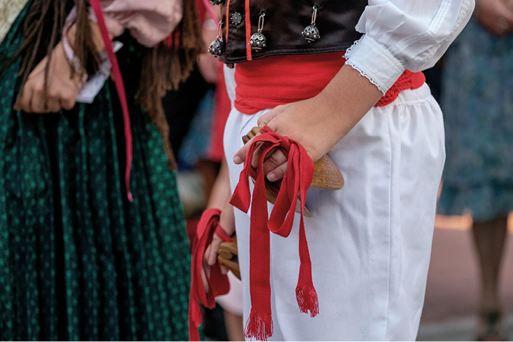 Traditionelle Tracht von Ibiza mit weissem Hemd, rotem Gürtel und verziertem dunklem Westen; die Person hält einen mit roten Bändern geschmückten Holzstab, im Hintergrund weitere Personen in folkloristischer Kleidung.