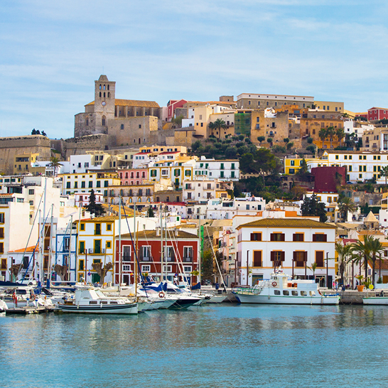 Bunter Hafen von Ibiza‑Stadt mit Booten und Yachten im ruhigen Wasser, farbenfrohen Häusern an der Uferpromenade und der historischen Kirche auf dem Hügel im Hintergrund.