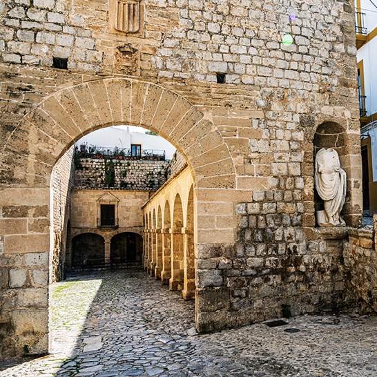 Steinerner Torbogen und historische Mauern der Altstadt von Dalt Vila, mit Kopfstatue in einer Nische und gepflastertem Durchgang im Sonnenlicht.