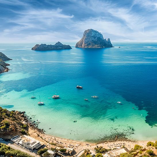 Blick auf die Bucht von Cala d’Hort mit türkisfarbenem Wasser, Booten und den markanten Felsen von Es Vedrà im Hintergrund unter hellem Küstenhimmel.