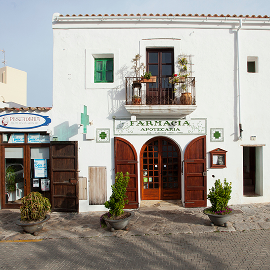Weiss getünchtes Dorfhaus in Sant Joan mit Apotheke und Fischladen, Holztüren, Balkon mit Blumen und traditioneller mediterraner Architektur an einer gepflasterten Gasse.
