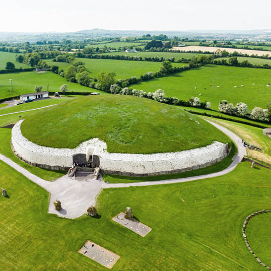 Newgrange aus der Vogelperspektive mit grasbewachsenem Hügel, weisser Steinfassade und umliegenden Feldern.