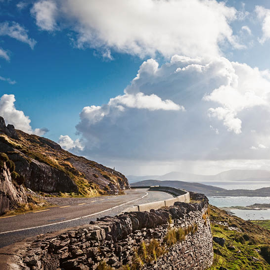 Küstenstrasse am Ring of Kerry mit Steinmauern, Felsen und Blick auf Meer und Hügel.