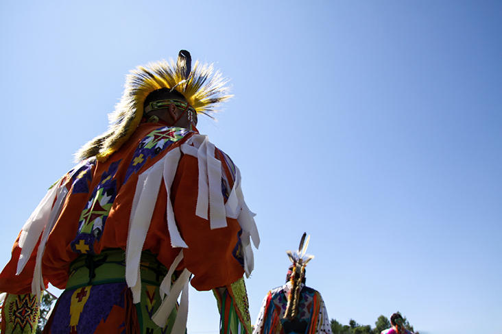 Zwei indigene Tänzer in traditioneller, farbenfroher Powwow‑Regalia unter blauem Himmel während einer kulturellen Feier.