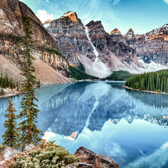 Klarer Bergsee im Banff National Park, umgeben von schneebedeckten Gipfeln und dichtem Nadelwald, mit perfekter Spiegelung im Wasser.