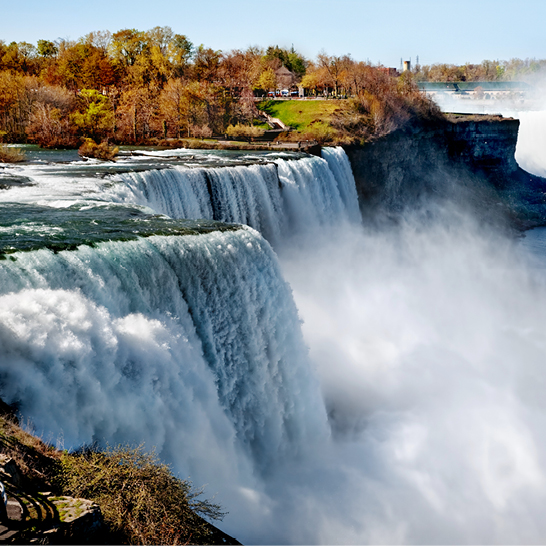 Breiter Wasserfall der Niagara Falls, umgeben von herbstlich gefärbten Bäumen, mit aufsteigendem Sprühnebel und Aussichtspunkt am Ufer.