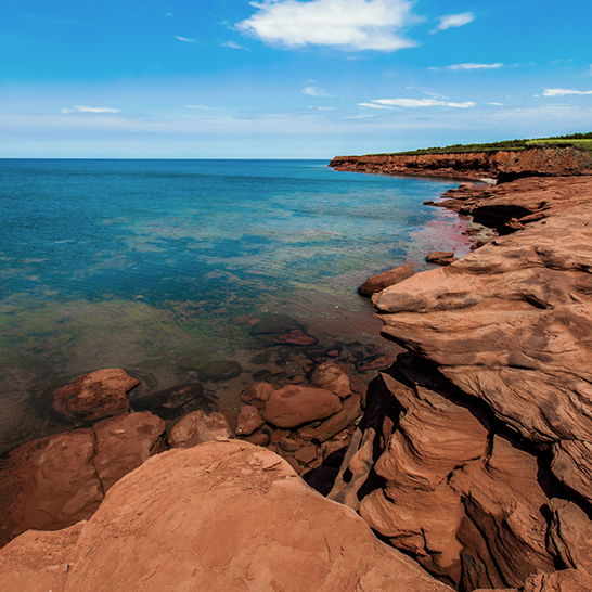 Rote Sandsteinklippen auf Prince Edward Island, umgeben von klarem blauem Meer und hellem Himmel mit vereinzelten Wolken.