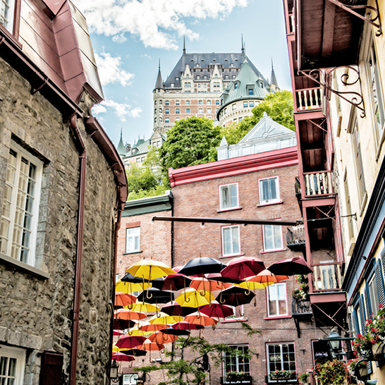 Bunte Regenschirme über der Rue du Petit‑Champlain in Québec City, flankiert von historischen Häusern, mit dem Château Frontenac auf dem Hügel im Hintergrund.