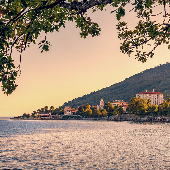 Küstenort in der Kvarner Bucht bei Sonnenuntergang mit ruhigem Wasser, Kirchturm und bewaldetem Hügel.