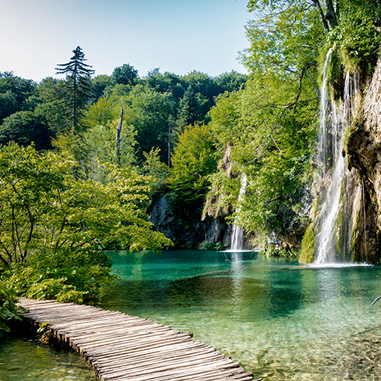 Holzsteg entlang eines türkisfarbenen Sees mit üppiger Vegetation und mehreren Wasserfällen im Nationalpark Plitvicer Seen.