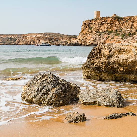 Sandstrand mit Felsen und sanften Wellen, im Hintergrund eine Klippe mit einem Wachturm bei Golden Bay, Malta.
