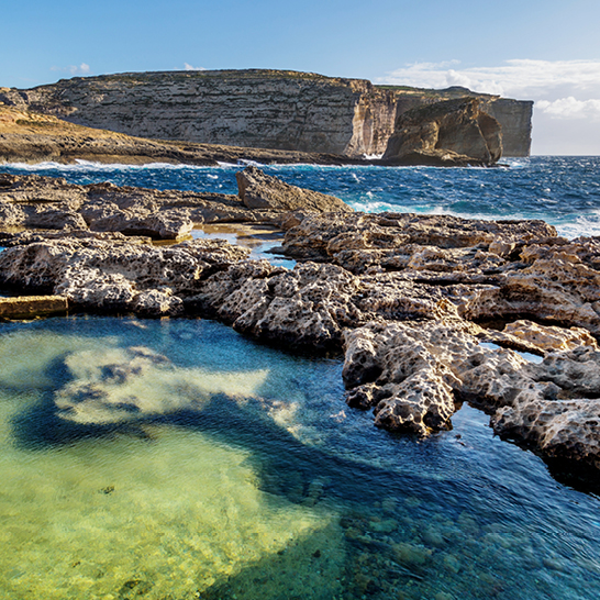 Rauhe Kalksteinfelsen und türkisfarbene Naturpools an der Küste von Gozo, Malta, mit steilen Klippen im Hintergrund.