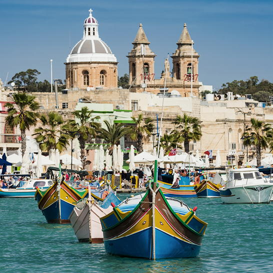 Bunte maltesische Fischerboote (Luzzus) im ruhigen Hafen von Marsaxlokk, mit Kirche und hellen Steingebäuden im Hintergrund.