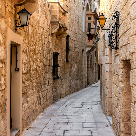 Schmale, gewundene Gasse mit hellen Steinmauern und Laternen in der Altstadt von Mdina (Medina), Malta.