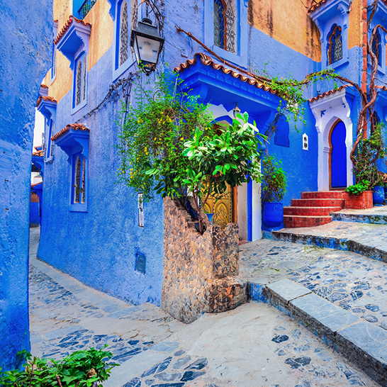 Enge blaue Gasse in Chefchaouen, mit Bögen, Pflanzen und roten Stufen zwischen traditionell gestrichenen Häusern.