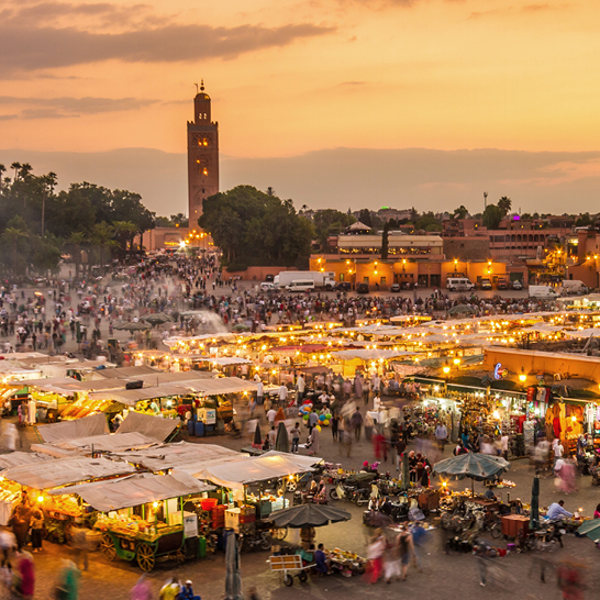 Abendstimmung auf dem Platz Djemaa el‑Fna in Marrakesch, mit beleuchteten Marktständen, Menschen und Minarett im Hintergrund.