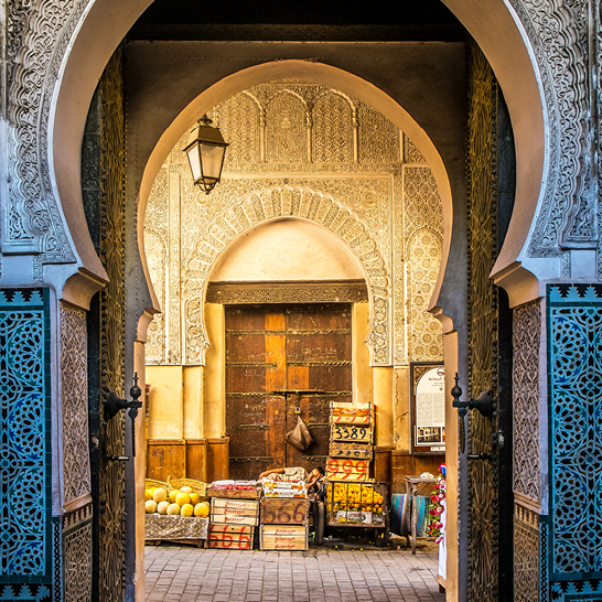 Verziertes Tor in der Medina von Fès, mit geschnitztem Bogen, Laterne und Obstkisten vor einer Holztür.