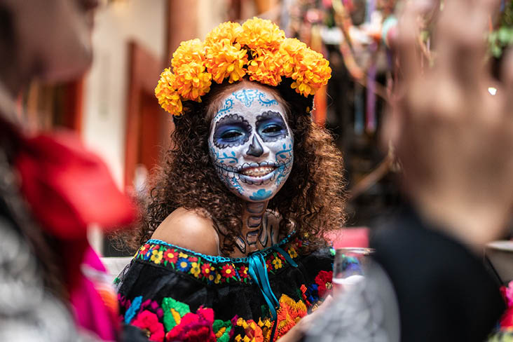 Person mit traditionellem Día‑de‑los‑Muertos‑Schminkdesign, orangefarbenem Ringelblumenkranz und bestickter schwarzer Kleidung in festlicher Umgebung.