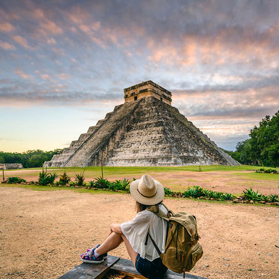 Person auf einer Bank mit Blick auf die Pyramide El Castillo in Chichén Itzá während eines farbenreichen Sonnenauf‑ oder ‑untergangs.