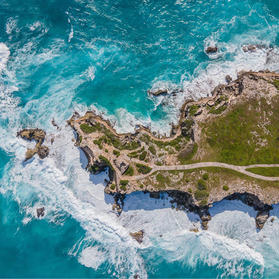 Luftaufnahme der Küste von Isla Mujeres mit felsigen Klippen, türkisfarbenem Meer und Wellen, die gegen das Ufer schlagen.