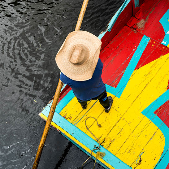 Person auf einem bunt bemalten Boot in Xochimilco, von oben gesehen, mit Strohhut und langem Stab über dunklem Wasser.