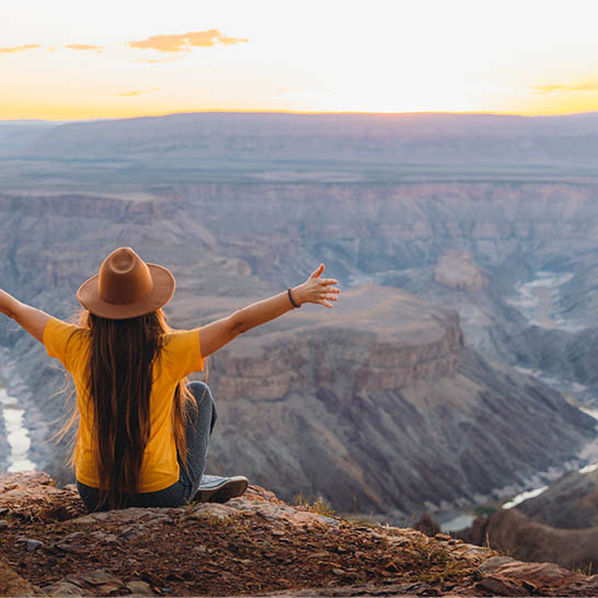 Person sitzt am Rand einer Felsklippe mit ausgebreiteten Armen und blickt über den weitläufigen Fish River Canyon im warmen Licht des Sonnenuntergangs.