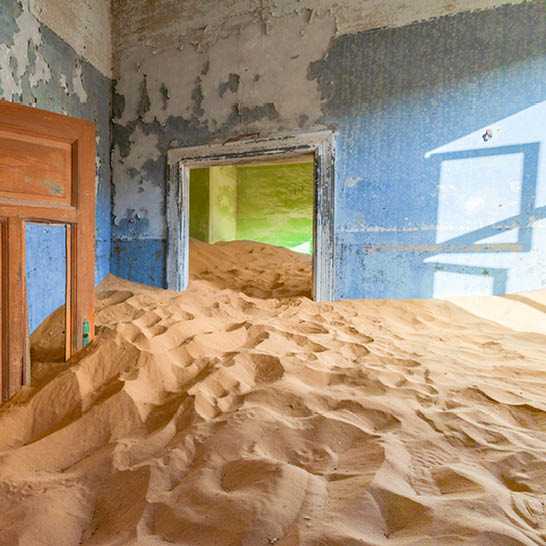 Verlassenes Zimmer in Kolmanskop, dessen Boden von hohen Sanddünen gefüllt ist; blau abgeblätterte Wände und Licht, das durch ein Fenster fällt.