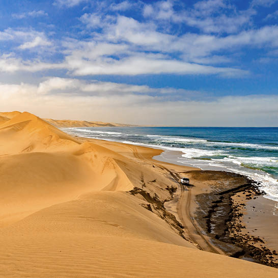 Hohe goldene Sanddünen treffen auf den Ozean an der Skeleton Coast, mit einem Fahrzeug auf einem schmalen Sandweg entlang der Küste.