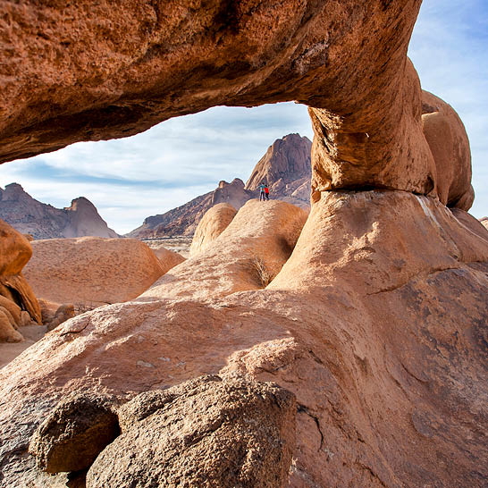 Natürlicher Felsbogen rahmt rötliche Granitformationen der Spitzkoppe, mit zwei kleinen Personen auf den Felsen unter teils bewölktem Himmel.