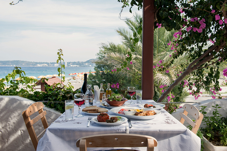 Table méditerranéenne avec plats grecs et vin, donnant sur la plage, la mer et les palmiers à Rhodes.