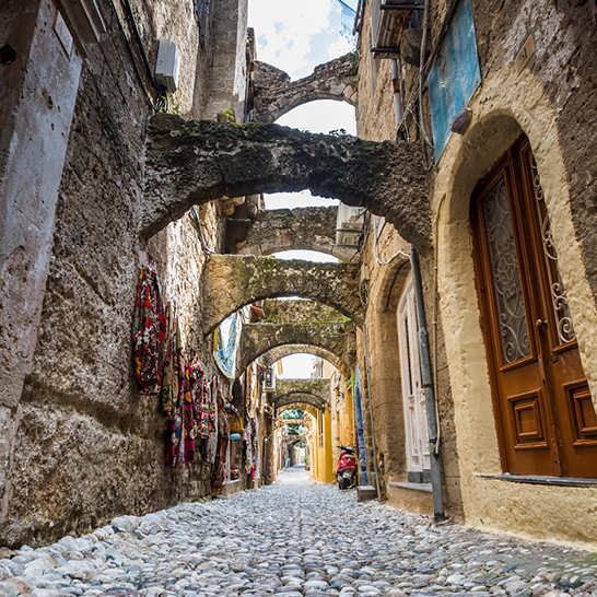Arc en pierre dans la vieille ville de Rhodes, avec murs patinés, verdure suspendue et ruelle pavée étroite.