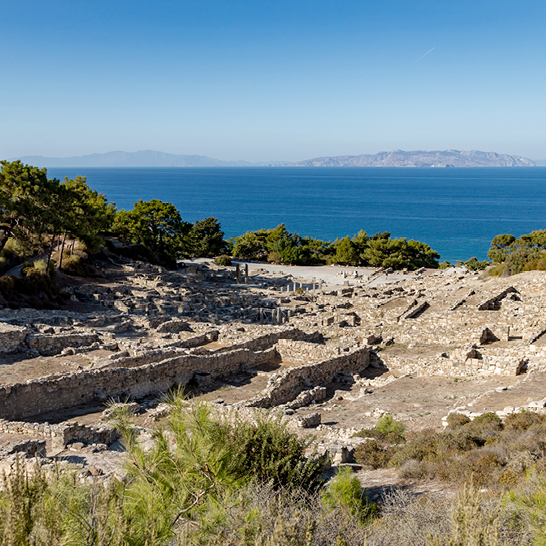 Ruines antiques de Kamiros à Rhodes, avec murs de pierre en terrasses surplombant la mer et la côte verdoyante.