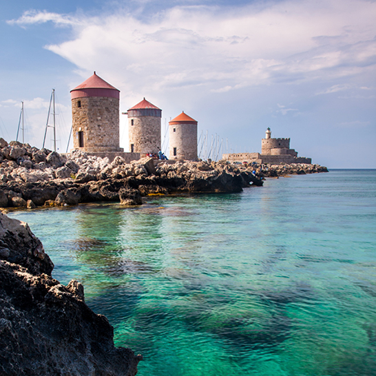 Moulins historiques du port de Mandraki à Rhodes, avec fort, phare et eau turquoise transparente.