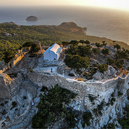 Vue aérienne du château de Monolithos à Rhodes, avec chapelle blanche perchée sur les rochers dominant la mer et les collines boisées.